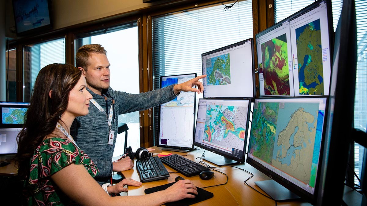 Två meteorologer på jour talar om kommande väder. Många monitor.